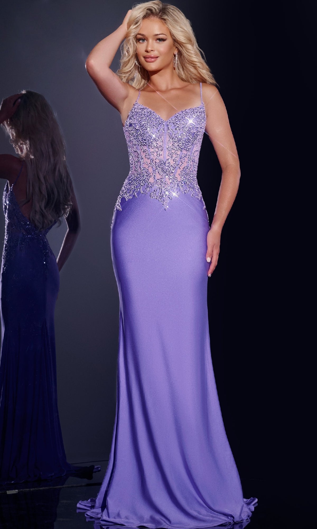 A woman stands against a dark background in the Jovani 42221 Sheer Beaded-Corset Long Prom Dress in violet, featuring spaghetti straps, floral lace details, and elegant beaded embroidery.